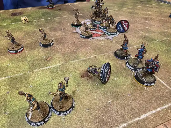 Two teams mid-game on a pitch with dice and a knocked-over base showing its underside